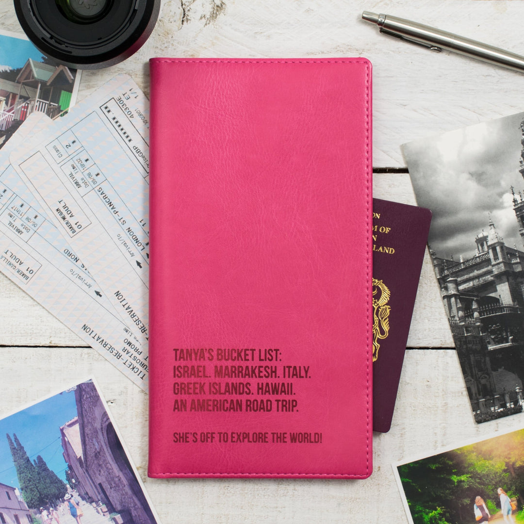 A pink personalized travel wallet with destination names embossed on it, placed on a wooden surface alongside passports, boarding passes, travel photos, and a pen.