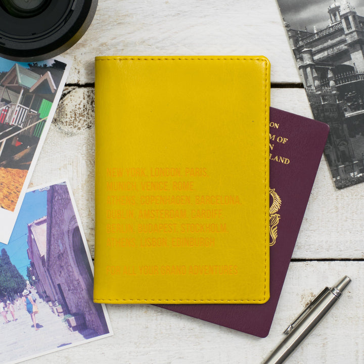 Personalized yellow passport holder featuring a list of famous cities, placed on a wooden table with travel documents and a pen.