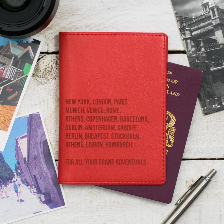 A red personalized travel wallet with city names embossed on it, placed on a wooden surface alongside passports, boarding passes, travel photos, and a pen.