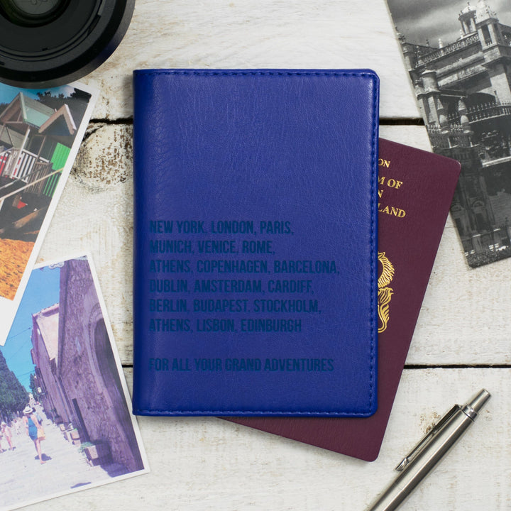 Royal Blue leather passport holder with embossed city names, surrounded by travel essentials like a passport, boarding passes, and postcards.
