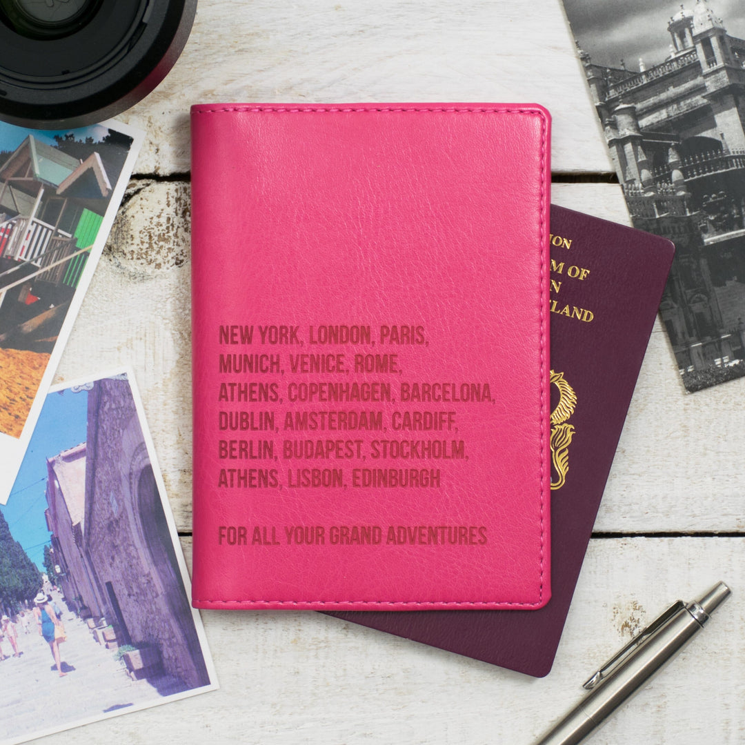 Personalized pink passport holder featuring a list of famous cities, placed on a wooden table with travel documents and a pen.