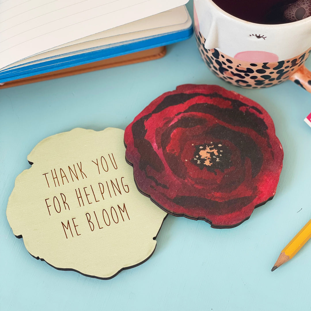  Two wooden coasters, one showcasing a vibrant red poppy and the other expressing gratitude with the message "Thank you for helping me bloom." The coasters are displayed on a desk alongside a mug and stationery, creating a cozy and appreciative scene.