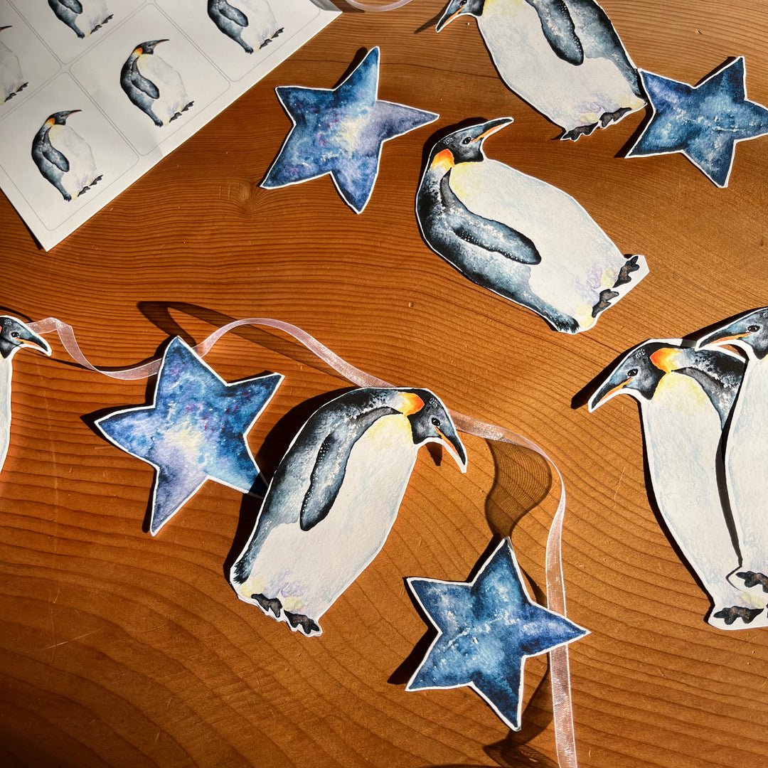 A penguin-themed garland with blue stars and penguins on a wooden table. The penguins are painted in watercolor and appear to be standing on a sheet of paper.