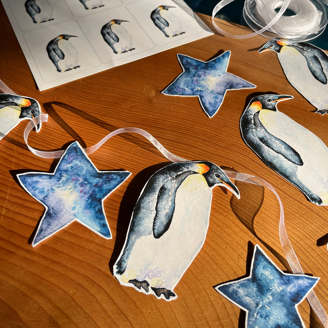 A penguin-themed garland with blue stars and penguins on a wooden table. The penguins are painted in watercolor and appear to be standing on a sheet of paper.