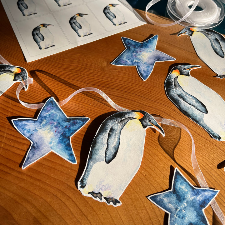 A penguin-themed garland with blue stars and penguins on a wooden table. The penguins are painted in watercolor and appear to be standing on a sheet of paper.