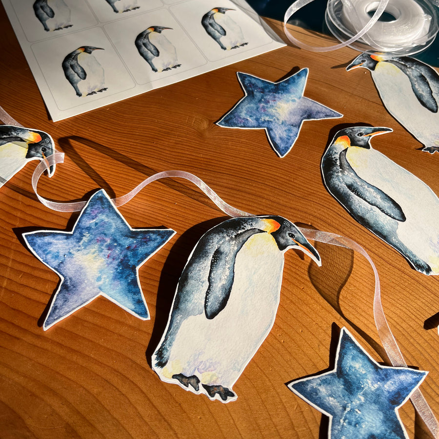 A penguin-themed garland with blue stars and penguins on a wooden table. The penguins are painted in watercolor and appear to be standing on a sheet of paper.