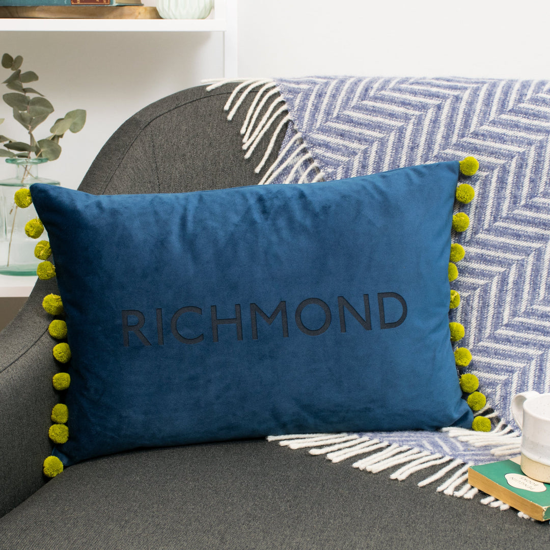 Richmond university graduation student gift, a blue pom pom cushion on a grey sofa against a white wall.