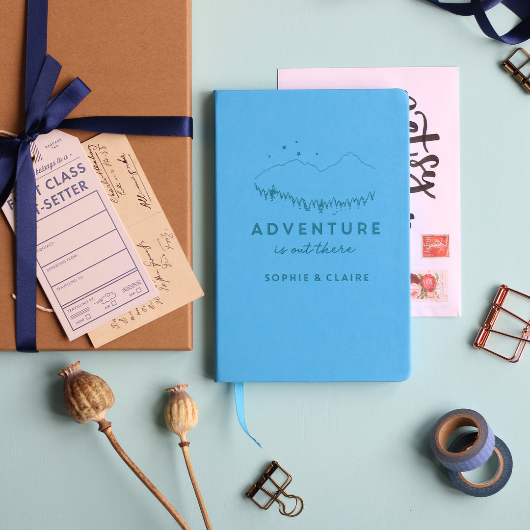 A top down image of a blue vegan leather notebook resting on a pale aqua table. The front of the notebook features an engraving of a mountain scene with stars and the text Adventure is out there. Personalised with the names Sophie & Claire. Surrounding the book are various stationary items.