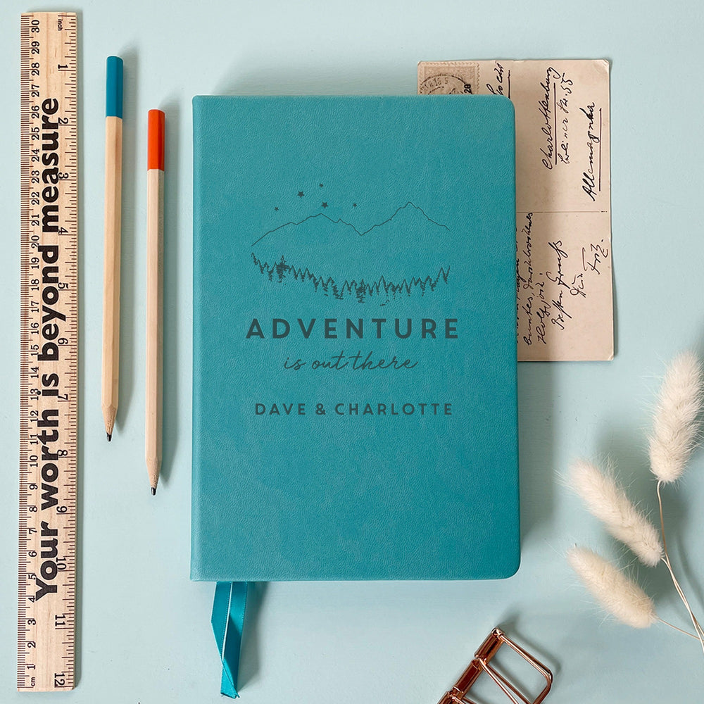 A top down image of a turquoise vegan leather notebook resting on a pale aqua table. The front of the notebook features an engraving of a mountain scene with stars and the text Adventure is out there. Personalised with the names Dave & Charlotte. Surrounding the book are various stationary items.