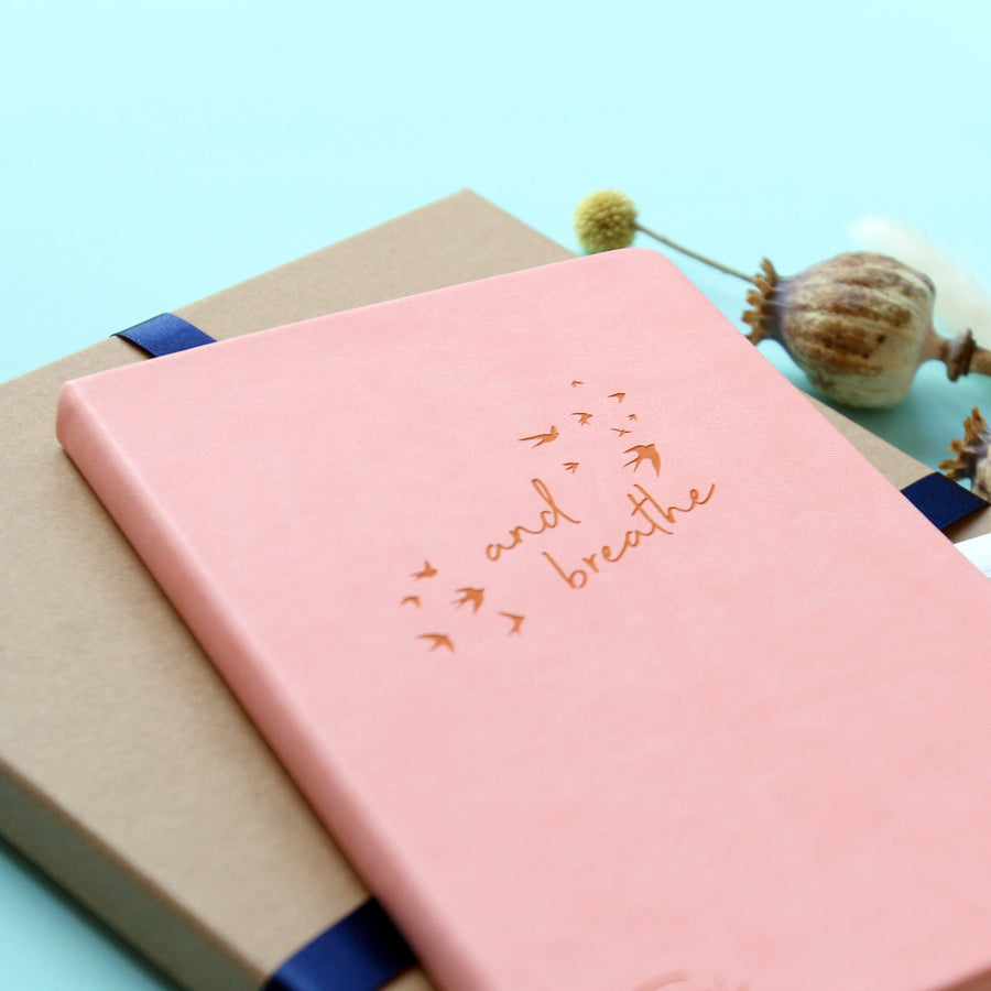 A top down image of a pink vegan leather notebook resting on a ribboned gift box. The front of the notebook features the words "and breathe" surrounded by birds. Surrounding the book are dried flowers.