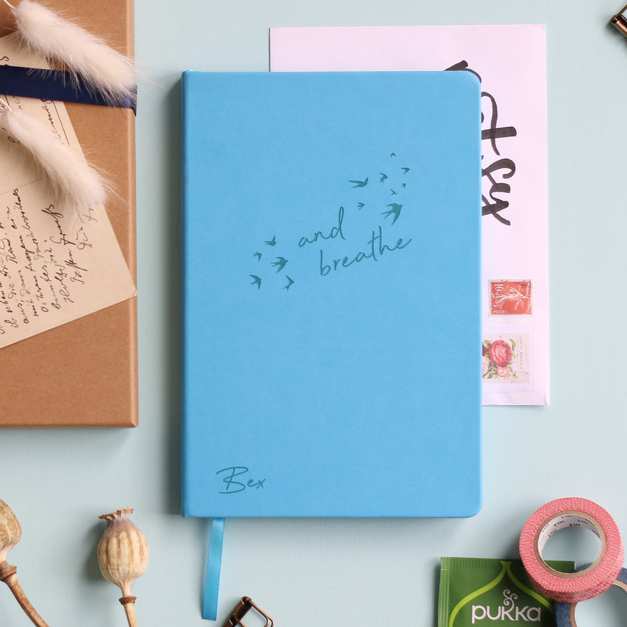 A top down image of a blue vegan leather notebook resting on a pale aqua table. The front of the notebook features the words "and breathe" surrounded by birds. The personalised engraving reads Bex. Surrounding the book are various stationary items and dried flowers.