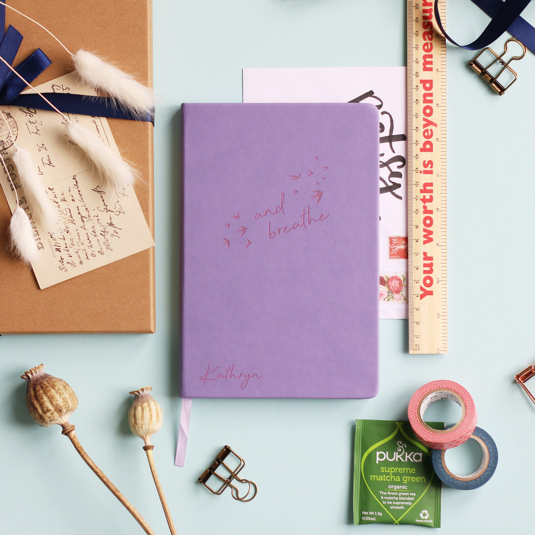 A top down image of a Lilac vegan leather notebook resting on a pale aqua table. The front of the notebook features the words "and breathe" surrounded by birds. The personalised engraving reads Kathryn. Surrounding the book are various stationary items and dried flowers.