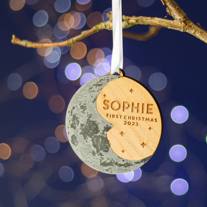 Wooden round decoration with a silver crescent moon and personalised text. Hanging on a branch in front of fairy lights. Perfect baby's first Christmas gift.