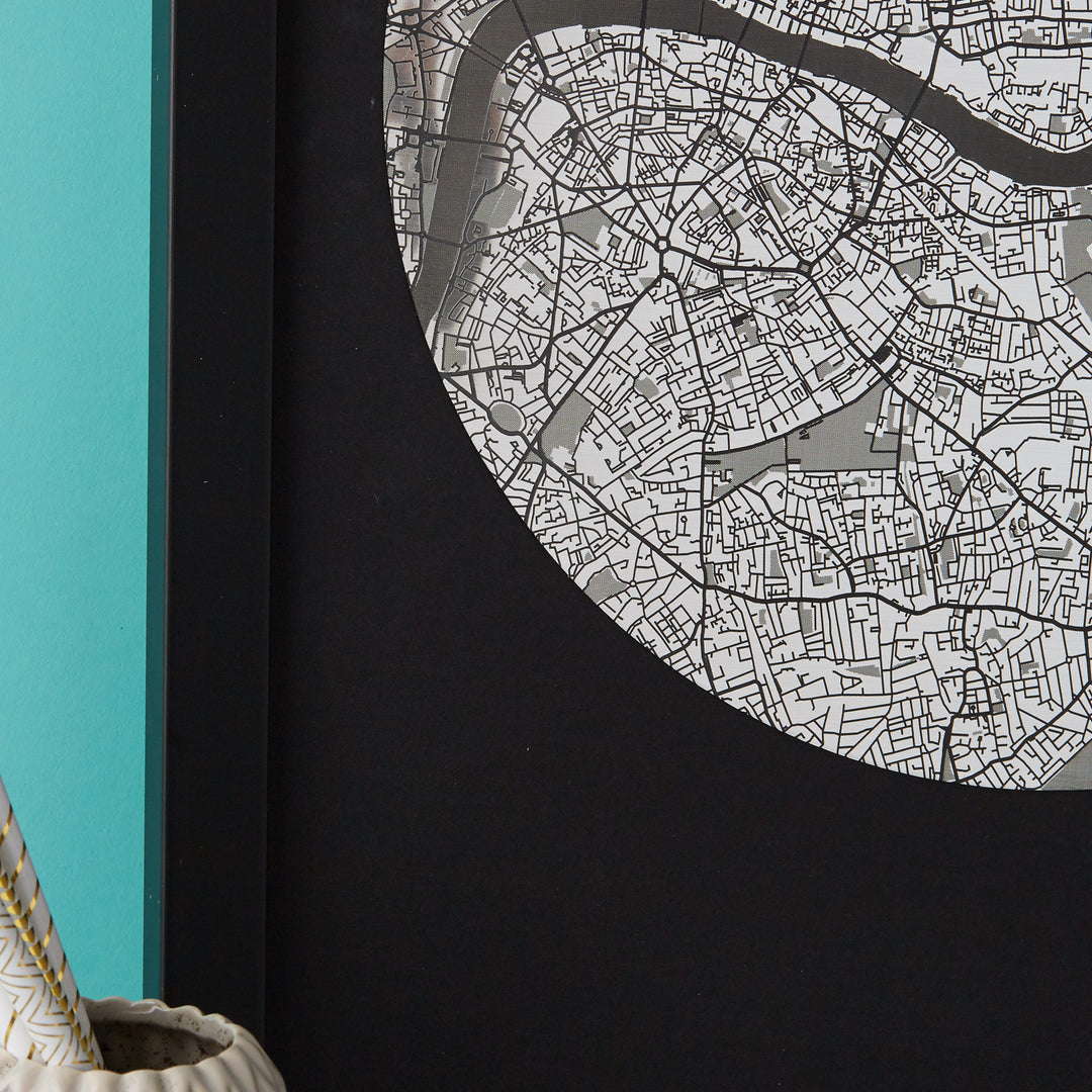 Close-up of a framed circular map of London in black and white, showing detailed street patterns. A portion of a teal wall and a light-colored object are visible in the lower left corner.