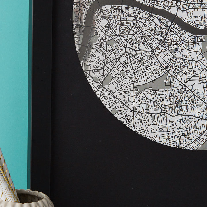 Close-up of a framed circular map of London in black and white, showing detailed street patterns. A portion of a teal wall and a light-colored object are visible in the lower left corner.
