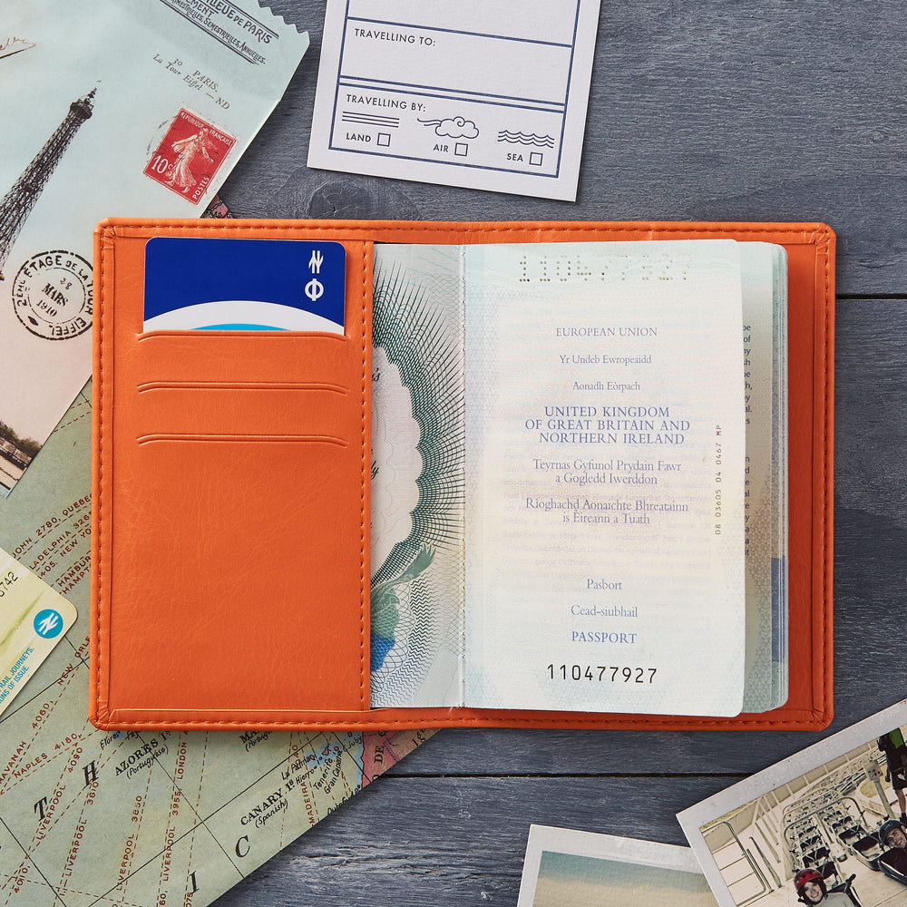 An open orange passport holder with a UK passport inside, laid on a table with vintage travel-themed papers and photos. The passport holder has a blue card in a slot and the passport shows the UK crest and text.