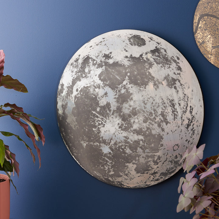A large metallic moon-themed wall plaque without text, mounted on a deep blue wall, surrounded by plants.