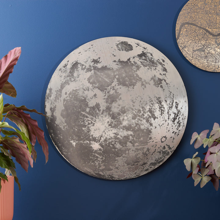 A round moon-themed wall plaque with a realistic lunar surface design, mounted on a deep blue wall, accompanied by plants and a metallic city map decor piece.