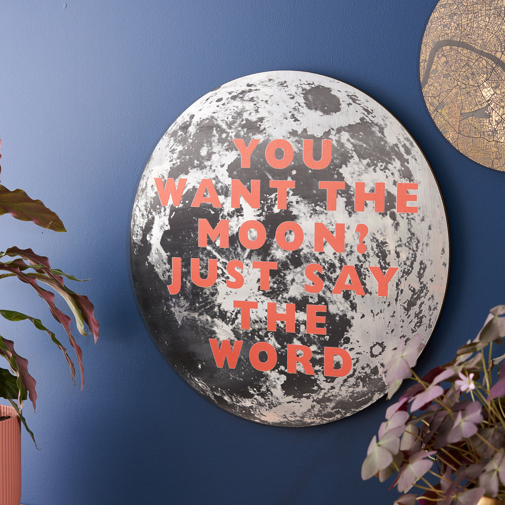 A large moon-themed wall plaque with the quote: "YOU WANT THE MOON? JUST SAY THE WORD" in bold red letters, mounted on a deep blue wall with surrounding decorative elements and plants.