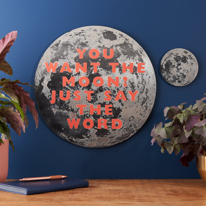 A large metallic moon-themed wall plaque with the quote "YOU WANT THE MOON? JUST SAY THE WORD" in bold orange letters, mounted on a deep blue wall with plants and a desk below.
