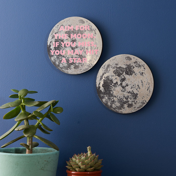Two moon-themed wall plaques on a dark blue wall. One has the quote, "AIM FOR THE MOON. IF YOU MISS, YOU MAY HIT A STAR," in pink text. There are also two potted succulents in the foreground.