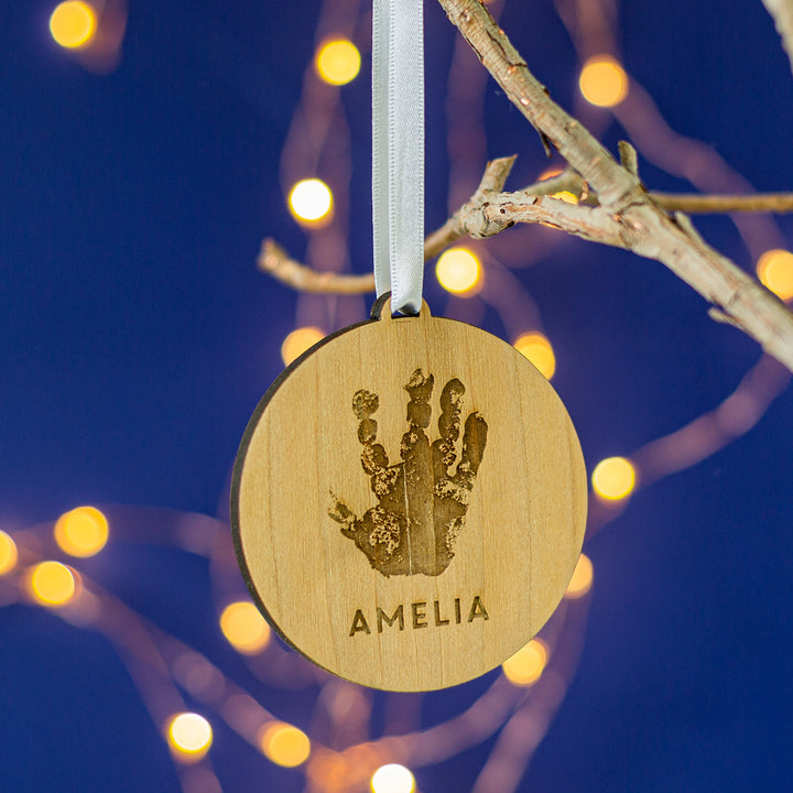 A personalized wooden Christmas tree bauble with a handprint design, hanging from a tree with lights and a name written on it.