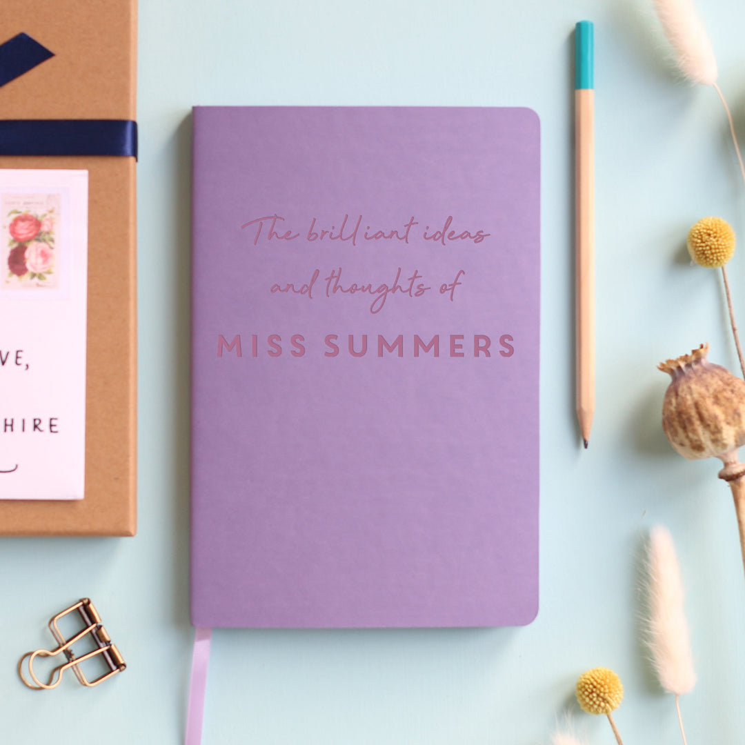 A top down image of a lilacvegan leather notebook resting on a pale aqua table. The front of the notebook is engraved with the phrase "The brilliant ideas and thoughts of" and is personalised with the name Miss Summers. Surrounding the book are various stationary items and dried flowers.