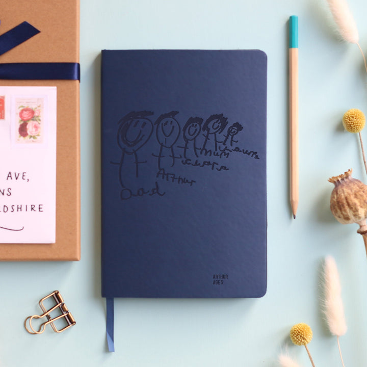 A top down image of a navy blue vegan leather notebook resting on a pale aqua table. The front of the notebook is personalised with an engraving of a child's drawing and the text Arthur age 5. Surrounding the book are various stationary items and dried flowers.