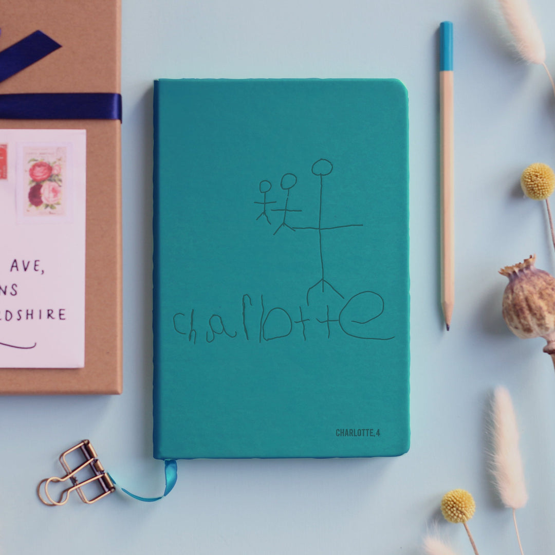 A top down image of a turquoise vegan leather notebook resting on a pale aqua table. The front of the notebook is personalised with an engraving of a child's drawing and the text Charlotte, 4. Surrounding the book are various stationary items and dried flowers.