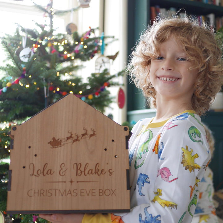 toddler holding a wooden house box showing the engravings of Santa's sleigh and the names Lola & Blake with a Christmas tree in the background.