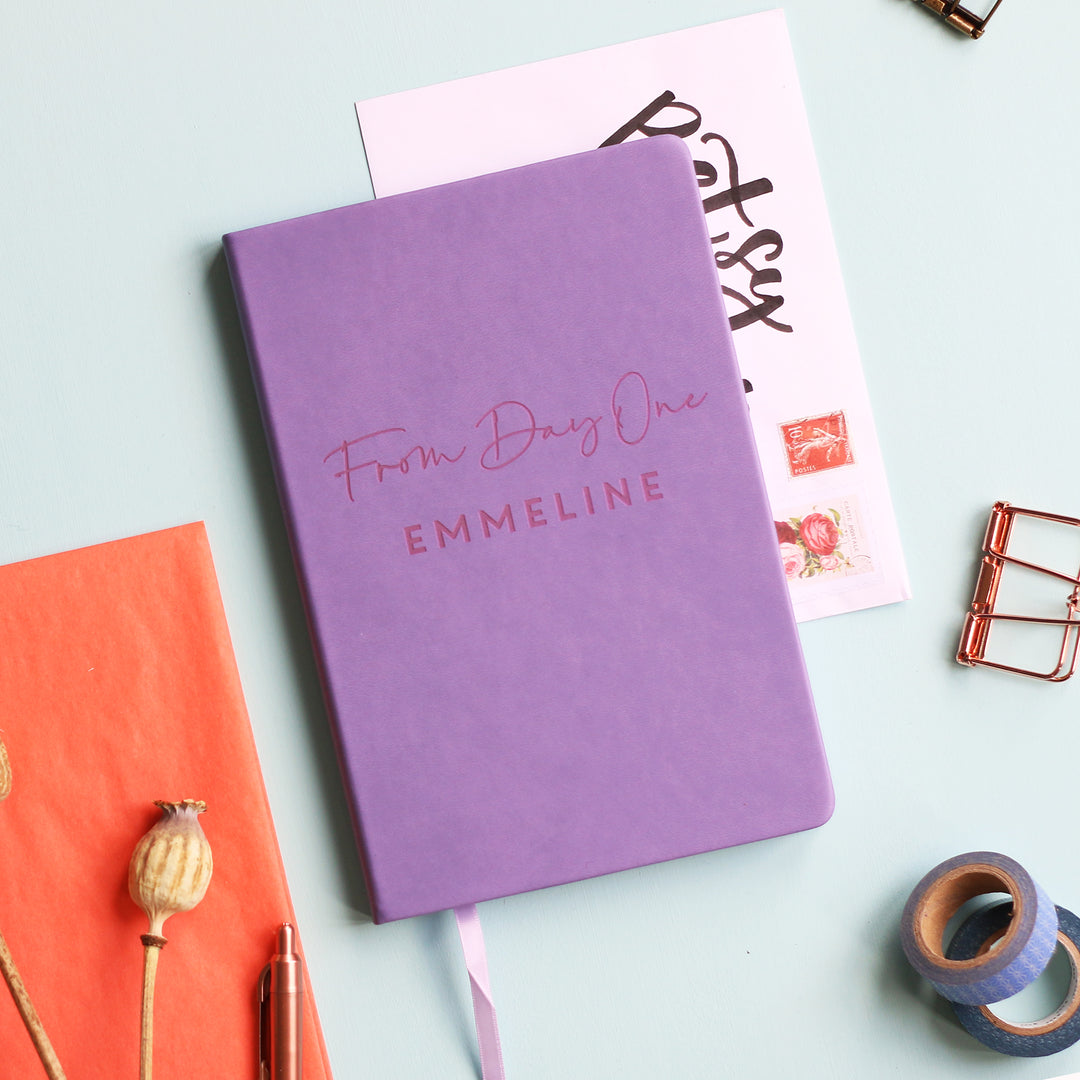 
A top down image of a lilac vegan leather notebook resting on a pale aqua table. The front of the notebook features the phrase "From Day One". It is personalised with the name Emmeline. Surrounding the book are various stationary items and dried flowers.
