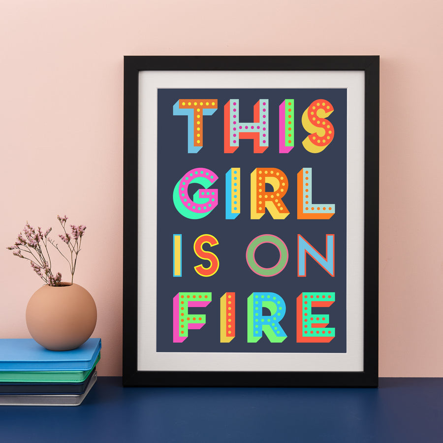 A framed print with the phrase "THIS GIRL IS ON FIRE" in colorful block letters against a dark background, sits on a blue surface. A small vase with twigs and a stack of books are next to it.