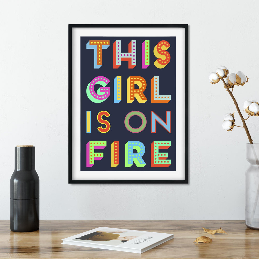 A framed print with the phrase "THIS GIRL IS ON FIRE" in colorful block letters on a dark background sits on a wooden table. A black vase, a cotton branch in a clear vase, and a book are also on the table.