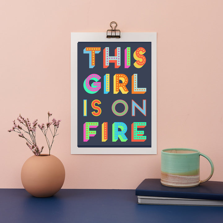 A framed print with the phrase "THIS GIRL IS ON FIRE" in colorful block letters on a dark background hangs on a pink wall. A vase with twigs, a book, and a mug are on a blue surface below.