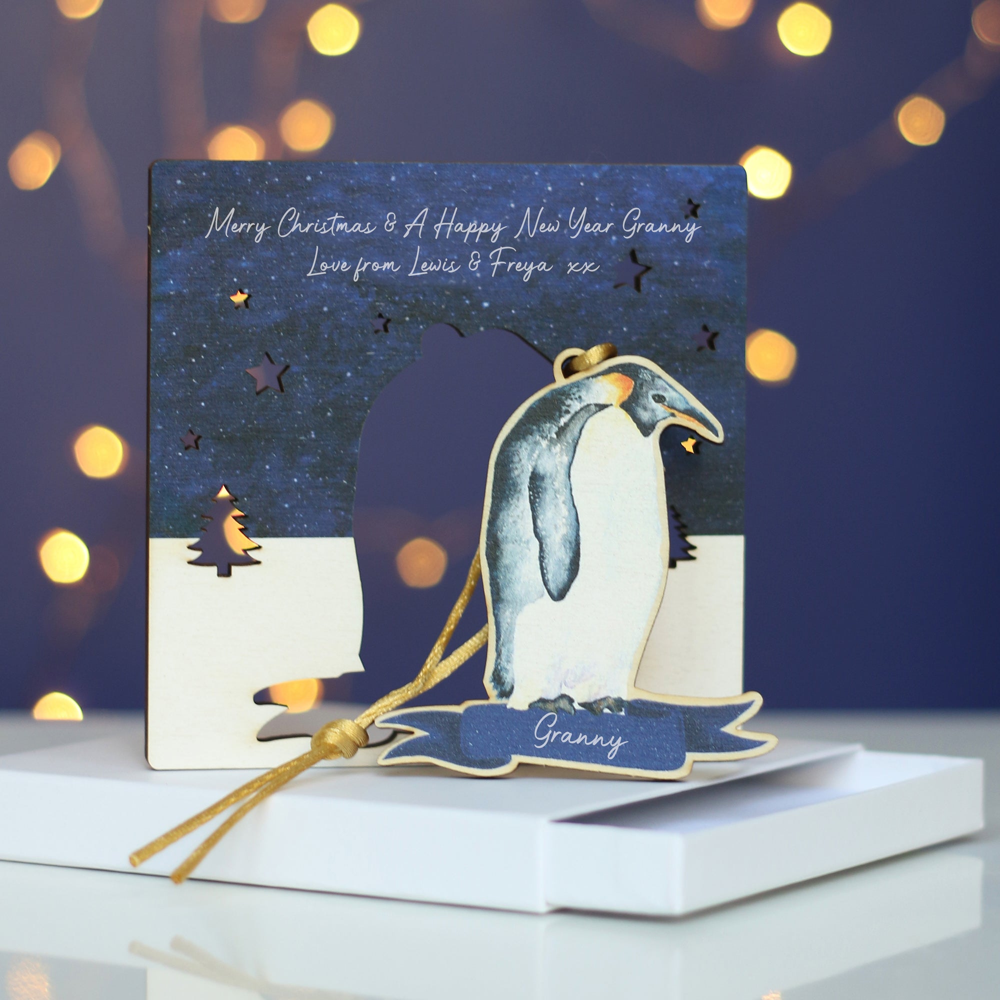 A wooden penguin decoration hangs from a branch. It is a watercolour of an Emperor penguin and has the name Granny at the bottom. In the background there's a blue wall and twinkly Christmas lights