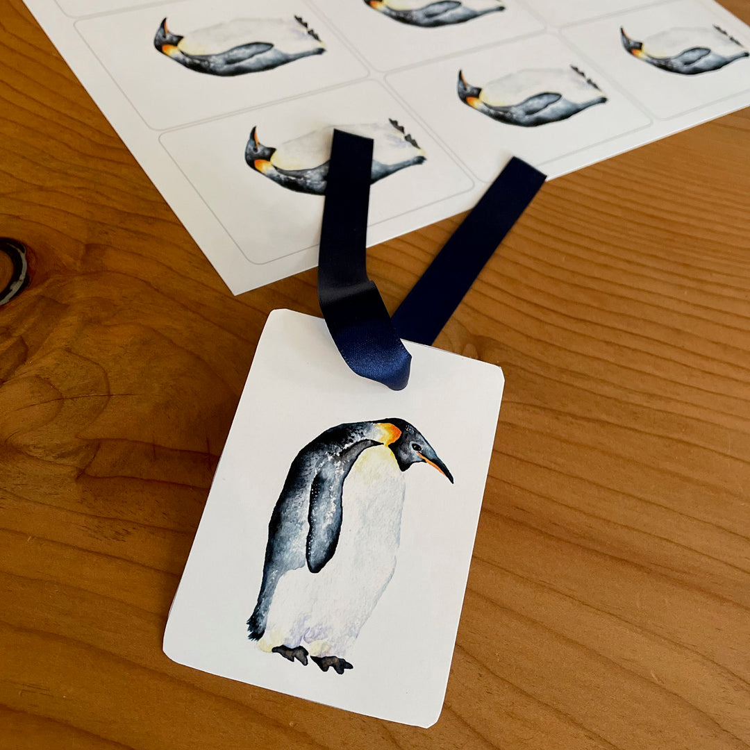 A close-up of a penguin gift tag with a blue ribbon. The tag features a watercolor painting of a penguin standing on a white background.