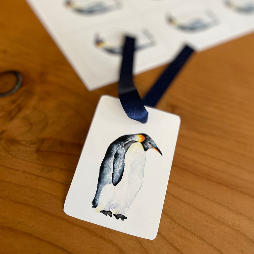 A close-up of a penguin gift tag with a blue ribbon. The tag features a watercolor painting of a penguin standing on a white background.