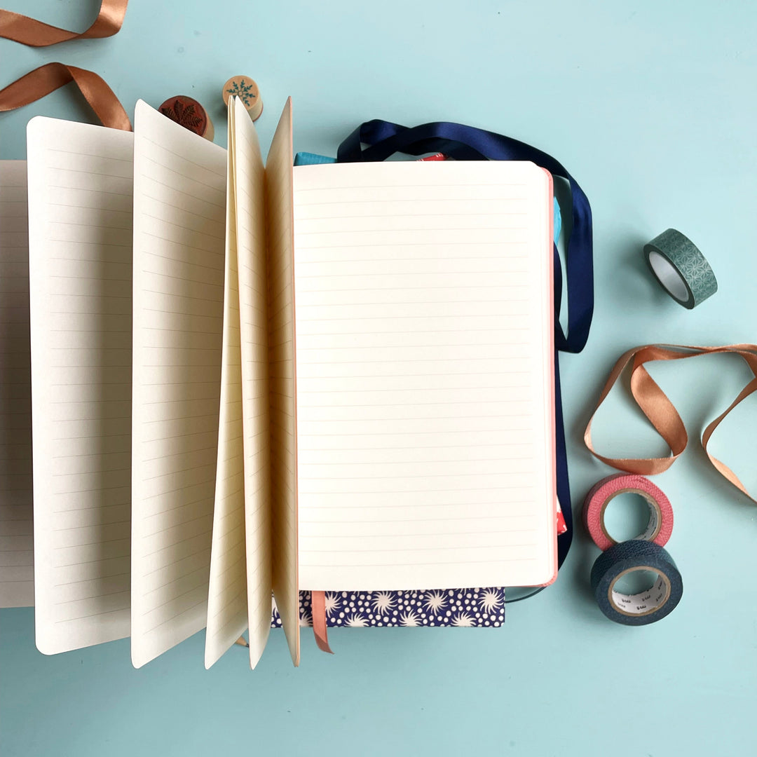 Personalised journal notebook open on pale aqua table to show lined pages and a matching colour ribbon place marker.  Washi tape, clips and other stationery items are scattered around it. 