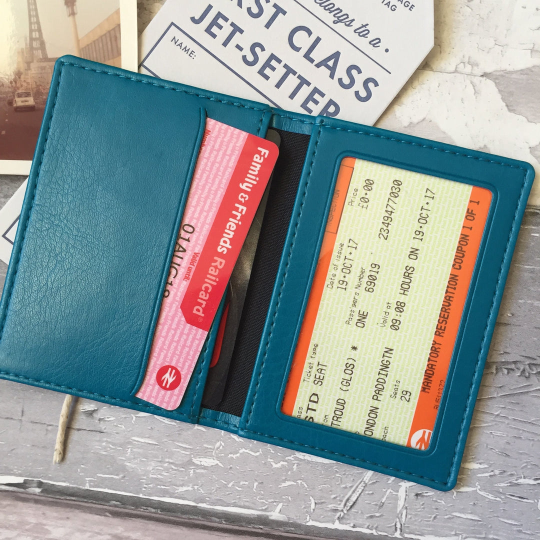 The turquoise leather cardholder minimalist wallet is lying open on a grey table. The interior shows a clear plastic window on one side where a photo or ID card can be displayed. On the other side, there are two credit card slots to hold cards, travel tickets, or a folded emergency banknote.