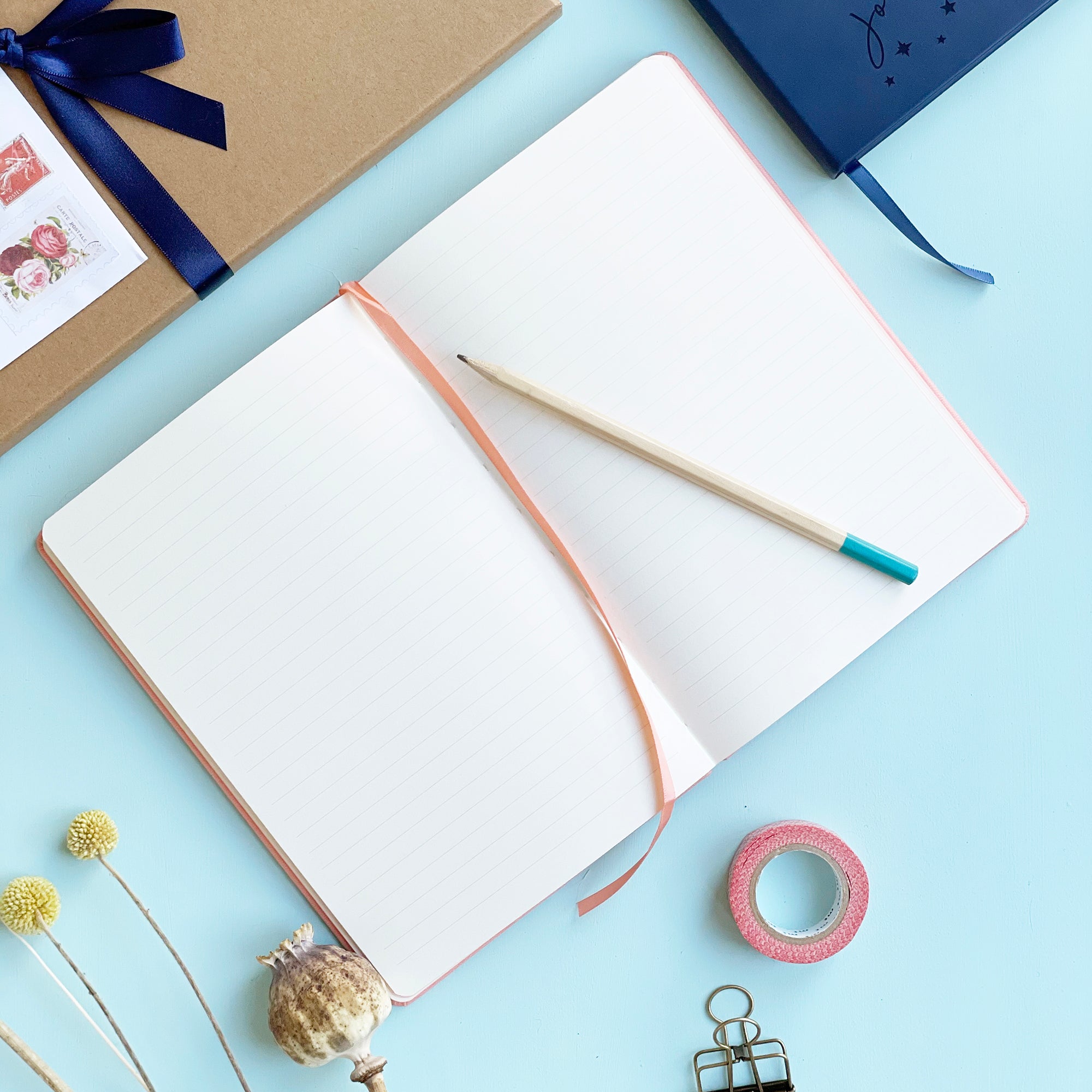 Personalised journal notebook lying flat open on pale aqua table to show lined pages and a matching colour ribbon place marker. A pencil is resting on the top of the notebook. Washi tape, clips and other stationery items are scattered around it. 