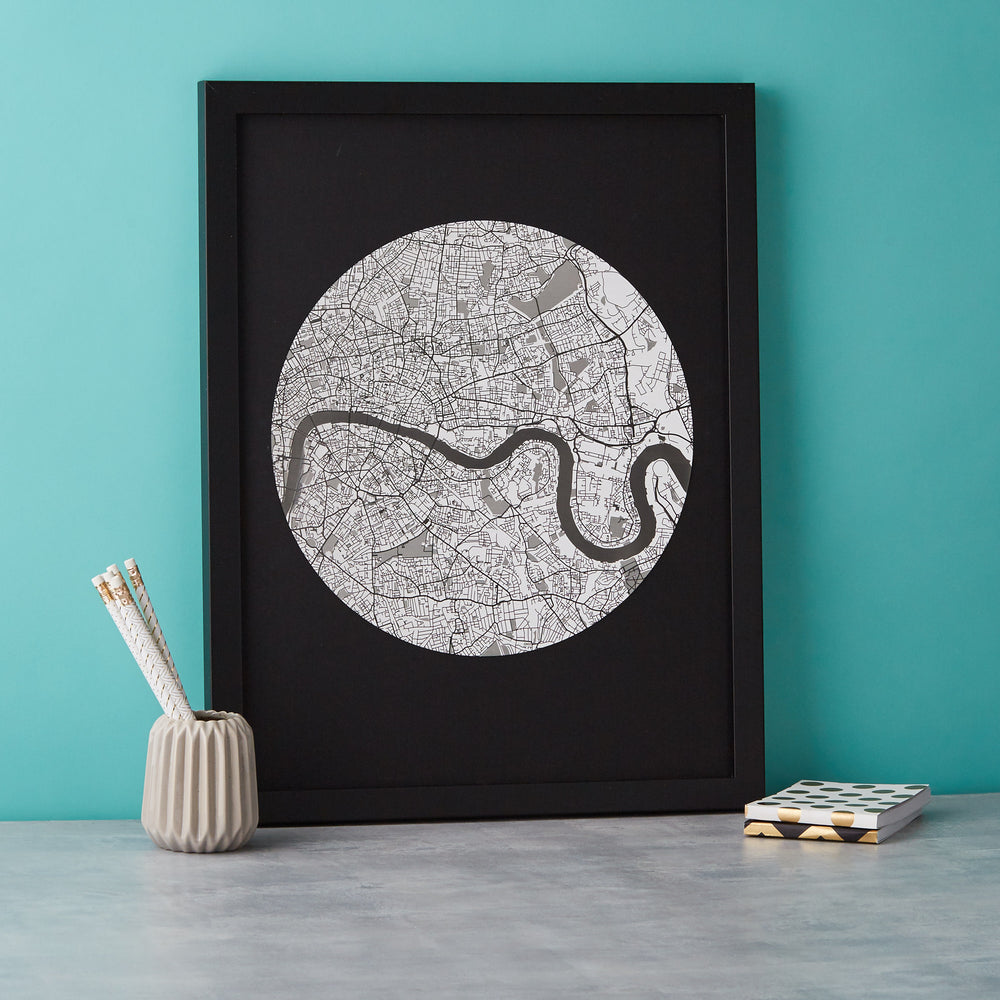  A framed circular map of London with the Thames River winding through it, displayed in black and white tones. Pencils in a gray holder and a small book are on the table next to the frame. The frame is black.