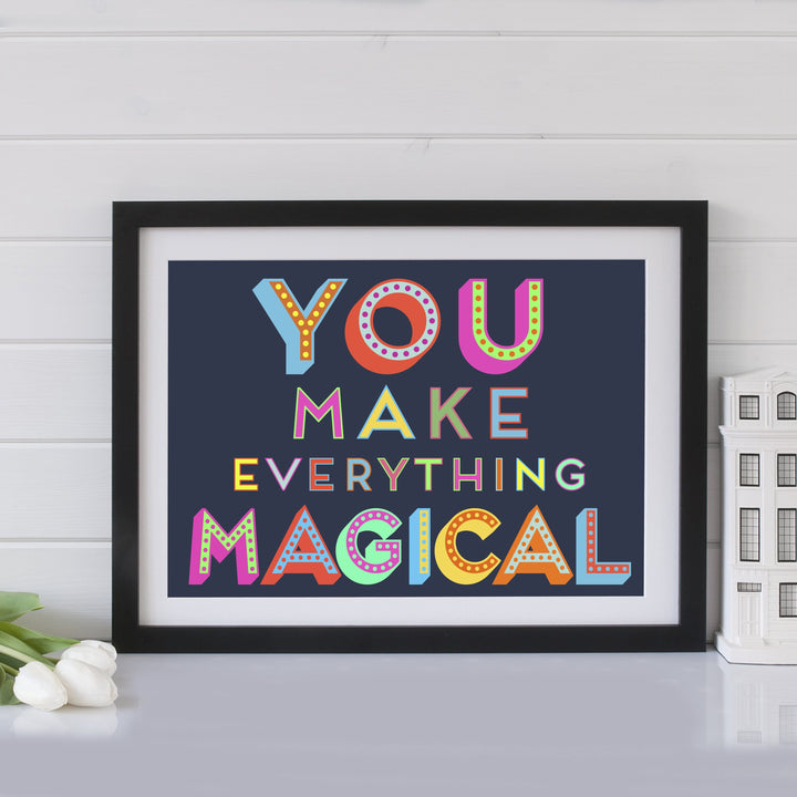 A vibrant and cheerful framed art print featuring the inspirational quote "You make everything magical" in a bold, colorful design. The artwork is displayed on a shelf with a vase of tulips and other decorative elements.