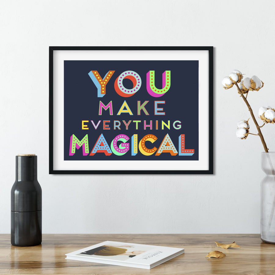 A framed art print featuring the inspirational quote "You make everything magical" in a bold, colorful design. The artwork is displayed on a wall with a vase of cotton flowers, a book, and other decorative elements.