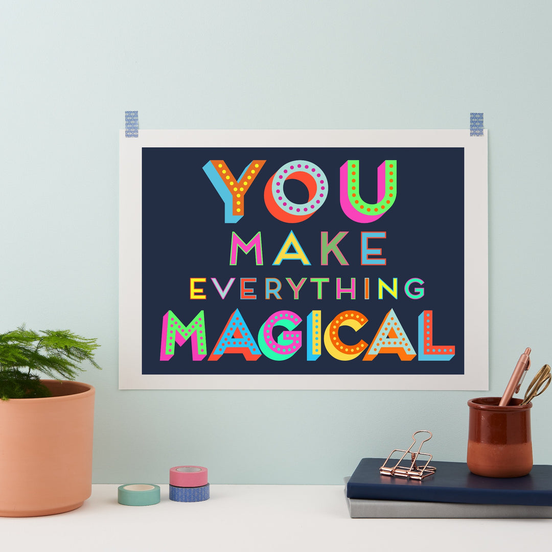 A framed art print with the motivational quote "You make everything magical" in colorful, playful typography. The artwork is displayed on a light blue wall with a plant, stationery, and other decorative elements.