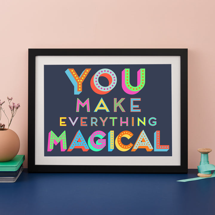 A vibrant and cheerful framed art print featuring the inspirational quote "You make everything magical" in a bold, colorful design. The artwork is displayed on a pink surface with a vase of flowers, a notebook, and other decorative elements.