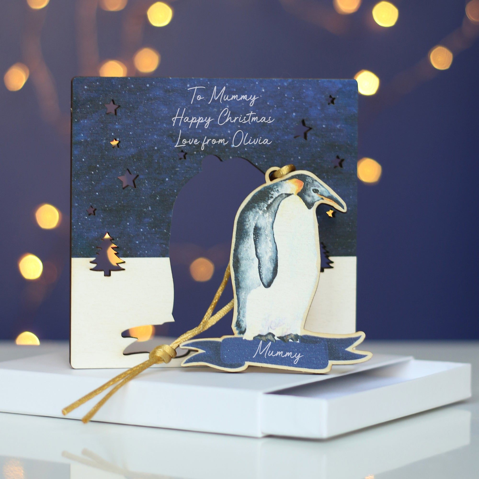 A wooden penguin Christmas decoration stands on a white gift box. The penguin has been "popped out" of a wooden surround that is also an alternative Christmas card. In the background there is a blue wall and twinkly lights 