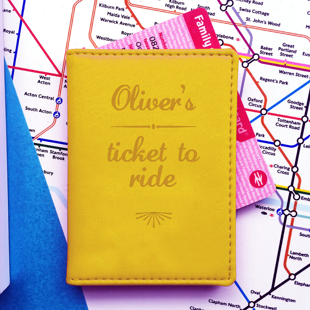 A yellow personalised vegan leather card holder is lying closed on top of a London Underground tube map. Poking out the side is a family railcard. The front of the travelcard cover is engraved with Oliver's Ticket to Ride