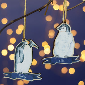 Two wooden penguin decorations hang from a branch. Watercolours of an Emperor penguin adult bird and a chick. They have the name Daddy or Ebba at the bottom. In the background there's a blue wall and twinkly Christmas lights