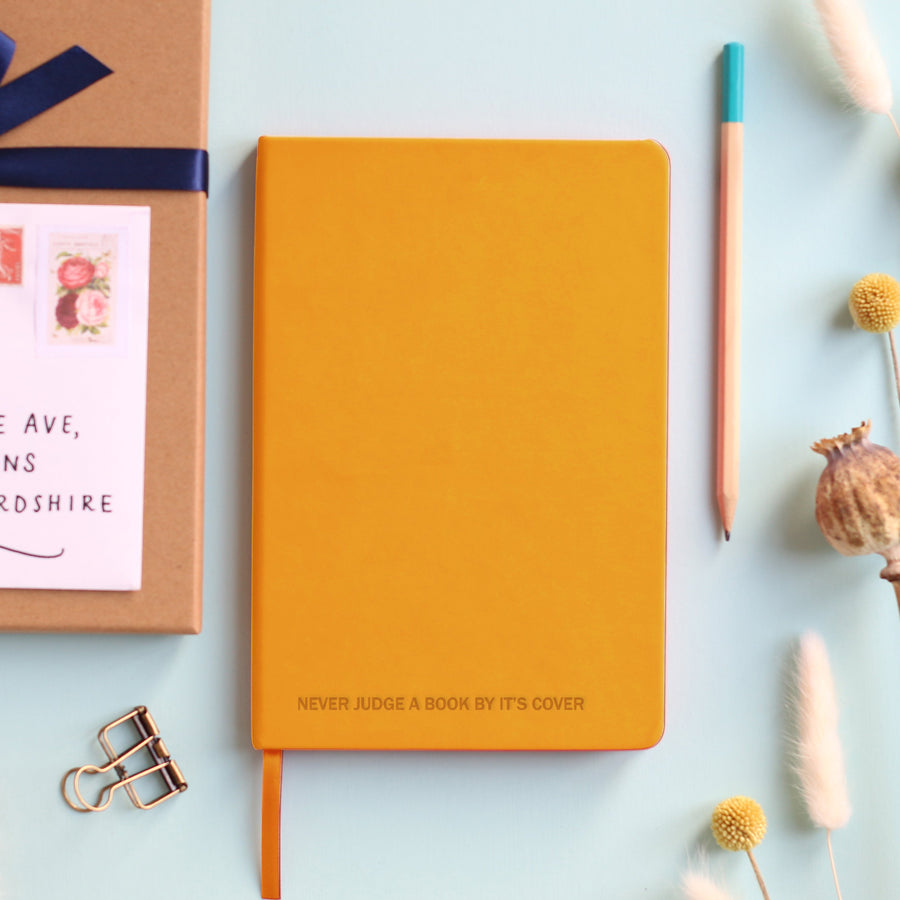 A top down image of a mustard yellow hardcover vegan leather notebook resting on a pale aqua table. The bottom of the book is engraved with a personalised message reading “Never judge a book by its cover.” Surrounding the book are various stationary items and dried flowers.