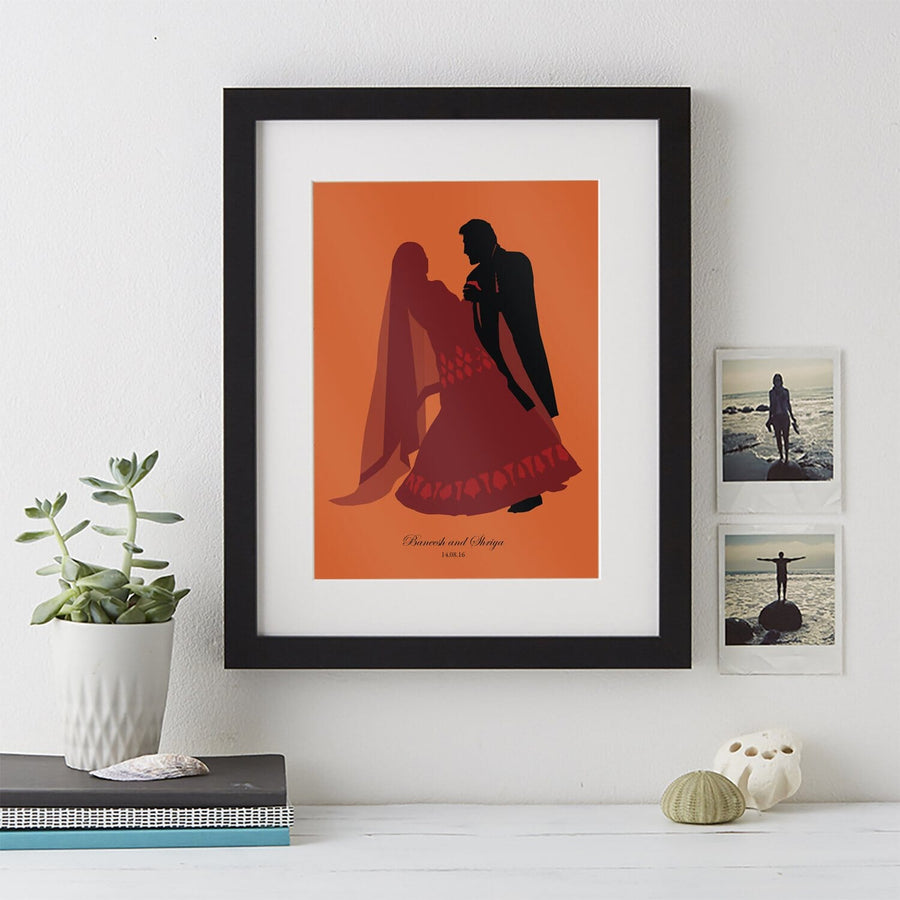 A framed silhouette portrait of a couple in traditional wedding attire on an orange background, displayed on a white wall with small photos and a potted plant nearby.